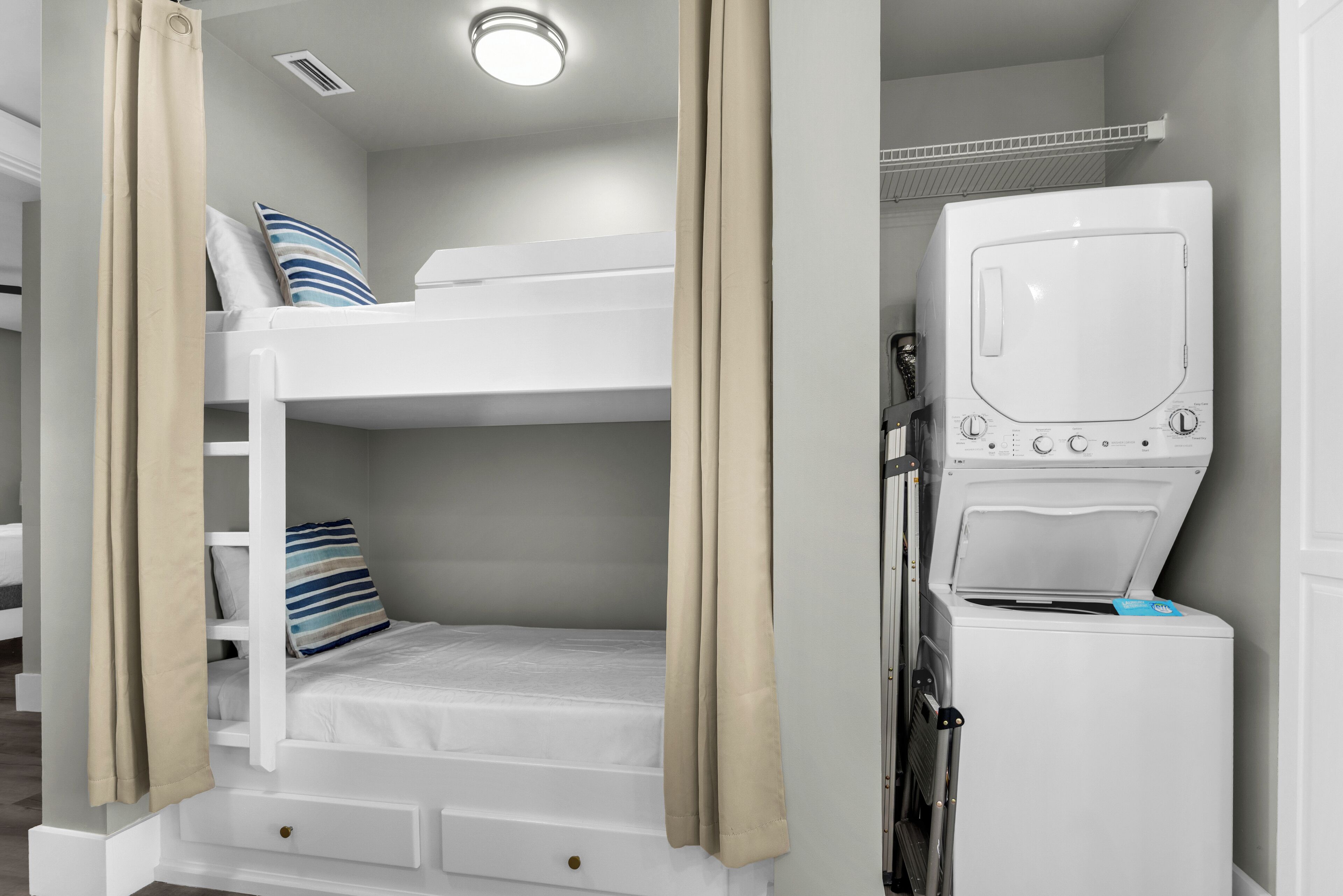 A bunk bed with a washing machine and a shelf unit in a room with beige curtains.