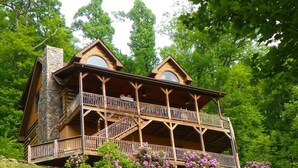 Eine zweistöckige Holzhütte mit einem Steinkamin, Holzbalkonen und einer üppigen Umgebung aus Grünpflanzen und violetten Blüten.