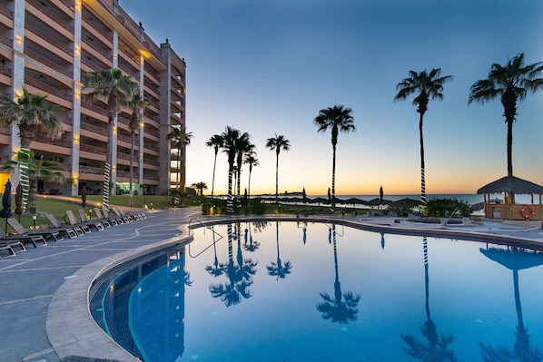A hotel pool area with a clear view of the pool, lounge chairs, palm trees, and a building with balconies.