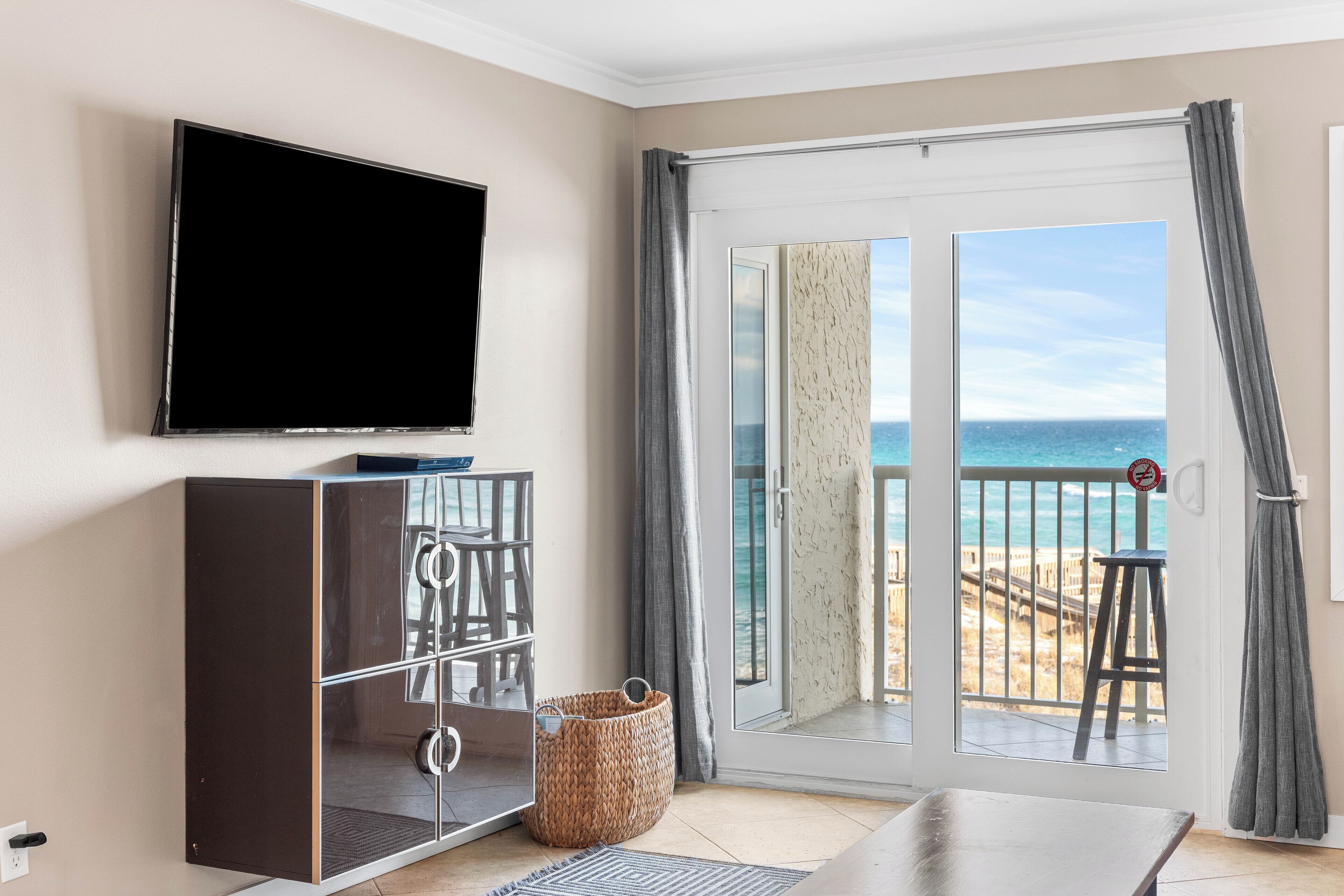 A room with a TV, a sliding glass door to the beach, a small table, and a wicker basket.