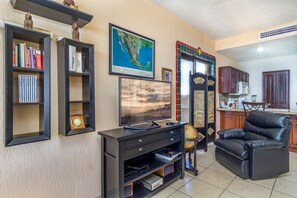 Sala de estar com televisão, estantes de livros, sofá de couro e área de cozinha ao fundo.