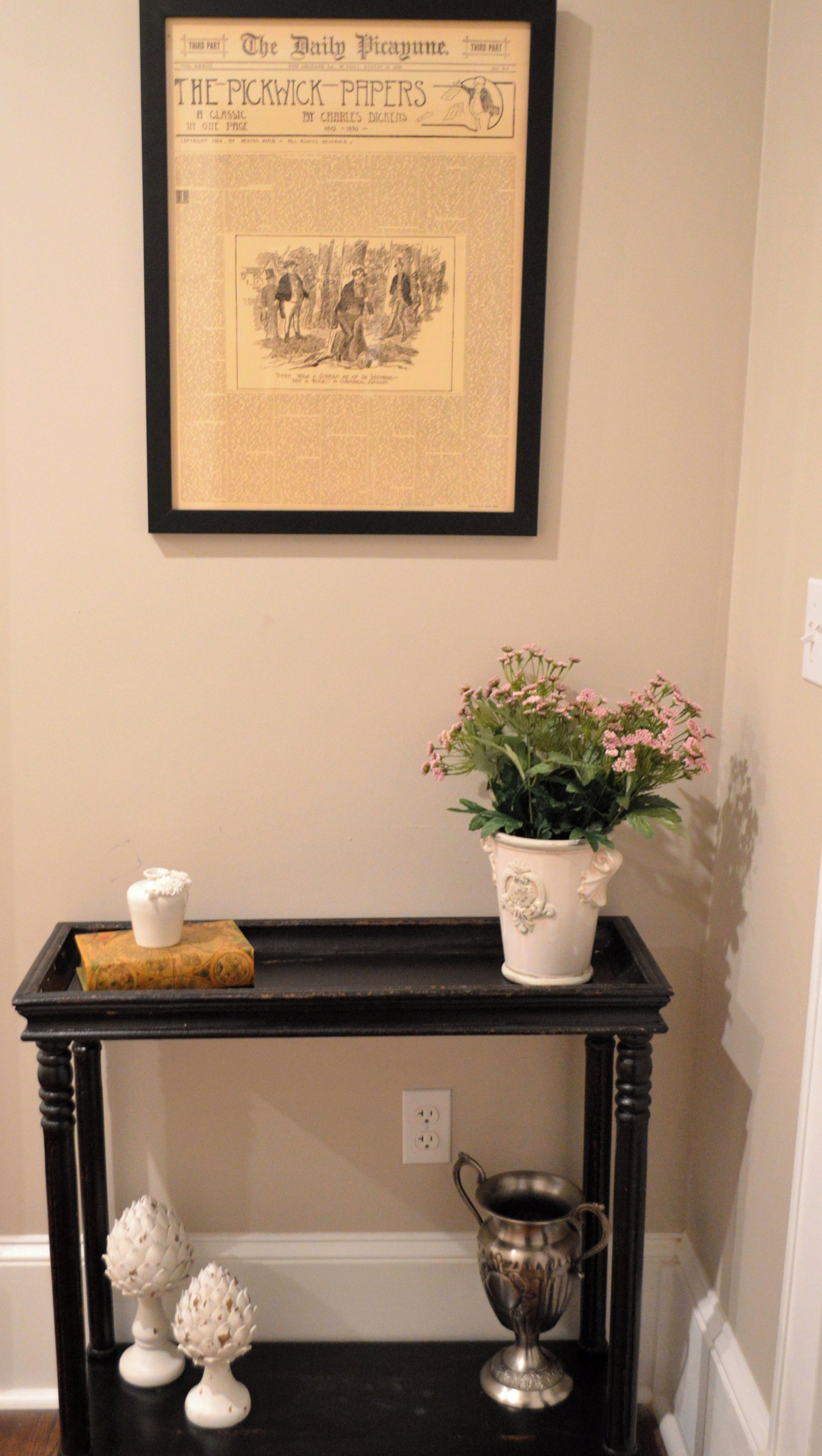 Un journal encadré accroché au mur, une petite table avec une plante, un objet décoratif et un pichet en argent.