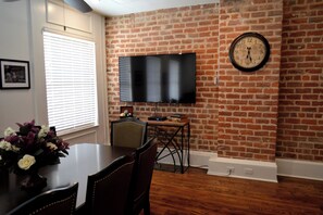 A dining area with a brick wall, a flat-screen TV mounted above a table, and a clock on the wall.