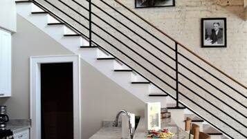 Une cuisine dotée d’un plan de travail en marbre, d’un évier et d’une table à manger en bois. Un escalier en métal noir mène à un palier où sont accrochées des photos encadrées.