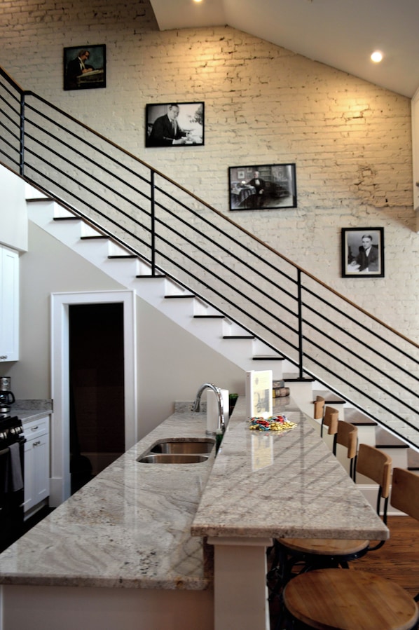 A kitchen with a marble countertop, a sink, and a wooden dining table. There are black metal stairs leading to a landing with framed pictures.
