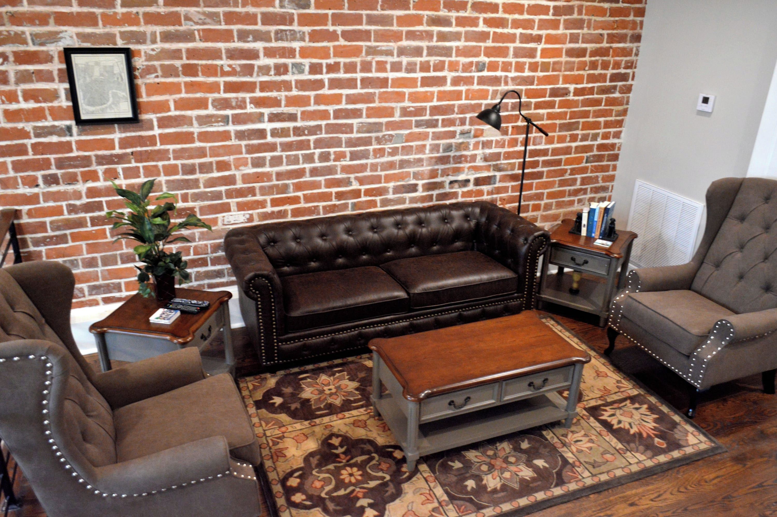 Un salon avec un mur en briques, un canapé en cuir marron, deux fauteuils, une table basse en bois et un tapis à motifs.