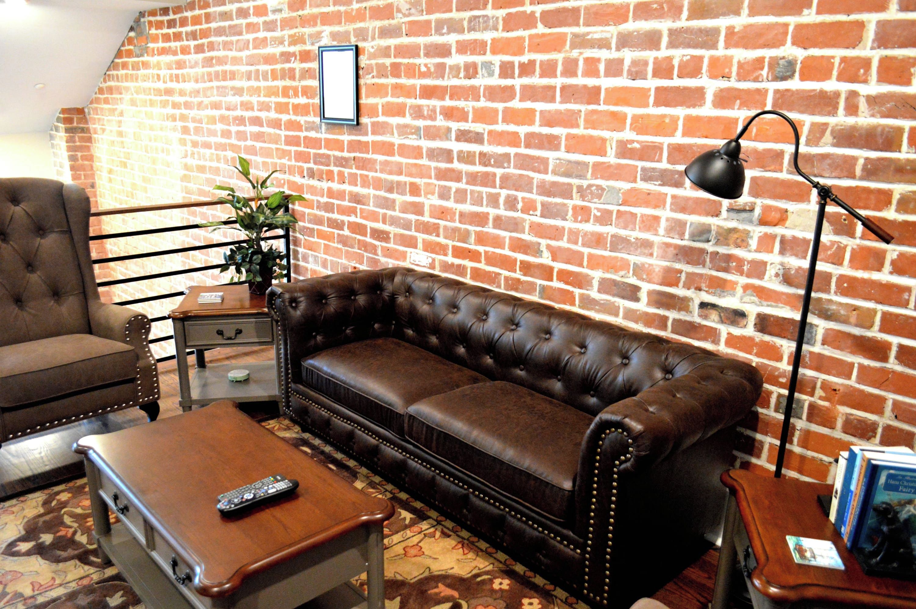 A living room with a brick wall, a leather sofa, a wooden coffee table, a side table, a potted plant, and a floor lamp.