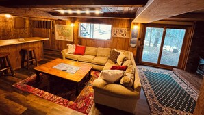 A cozy cabin interior with a wooden ceiling, a sofa, a coffee table, a bar area, and a large window offering a view of the outdoors. - Trailside Home on Alpen Trail with Hot Tub & Allows Dogs (Bessemer)