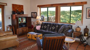 A living room with a television, sofa, and a view of a natural landscape through large windows.