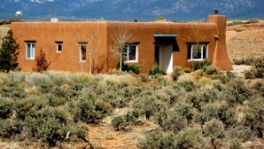 A single-story adobe building with a flat roof, surrounded by sparse vegetation and mountains in the background.
