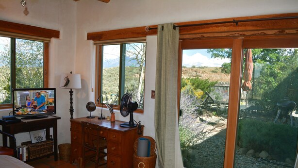 A room with a bed, a desk with a computer, a fan, and a view of a garden through large windows.