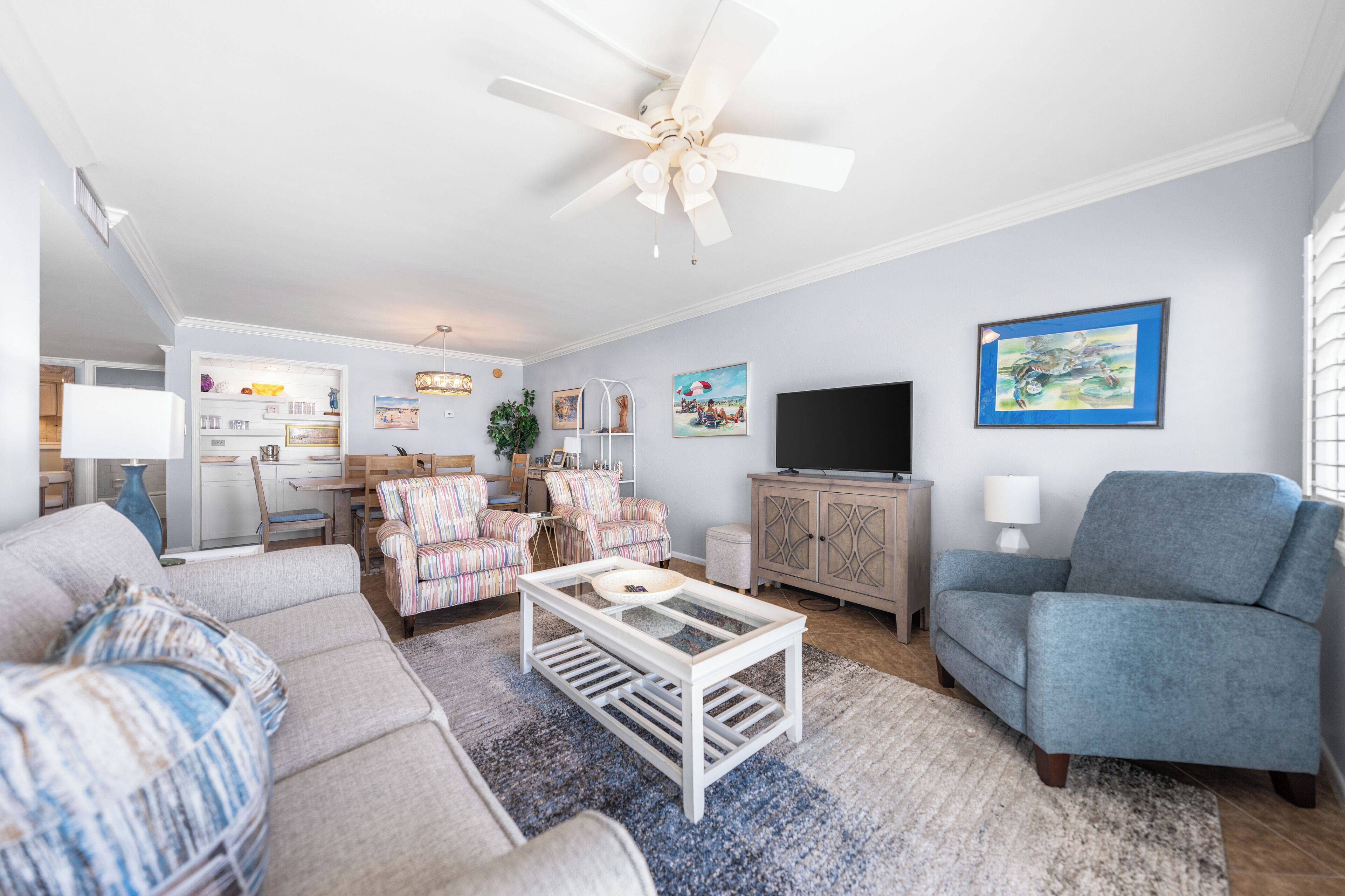 A living room with a sofa, armchair, coffee table, and TV. There is a ceiling fan, a cabinet, and a painting on the wall.