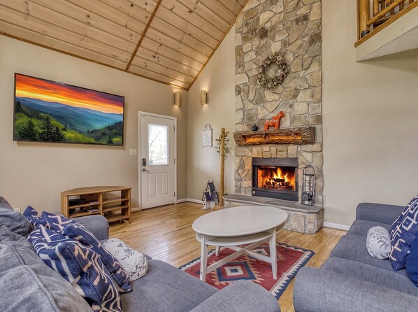 A living room with a stone fireplace, a painting of a sunset, and a cozy seating area. - Berg Stuga, Free Attraction Tickets, Game Room, Hot Tub, WIFI!!! (Townsend)