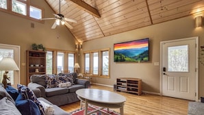 A spacious living room with a high ceiling, large windows, a gray sofa, a round coffee table, and a painting of a landscape.