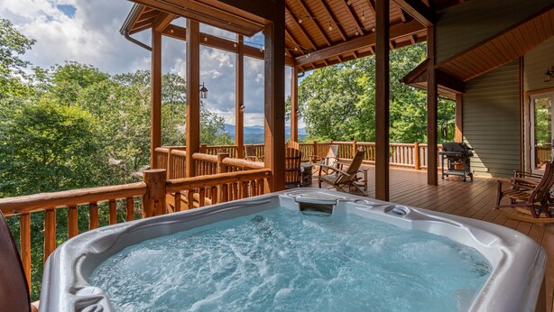 A wooden deck with a hot tub, outdoor furniture, and a view of trees and mountains.