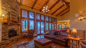 A spacious living room with a stone fireplace, leather sofas, large windows, and a wooden ceiling.