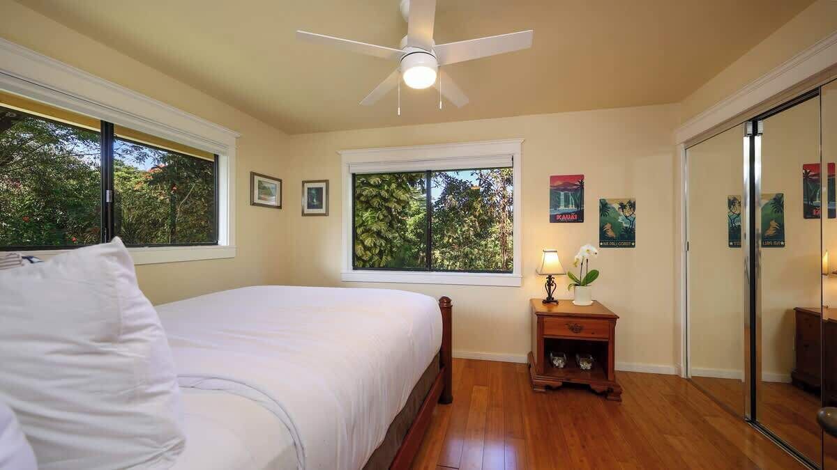 Une chambre avec un lit, un ventilateur de plafond, une fenêtre donnant sur des arbres et une table de chevet avec une lampe et une petite plante.