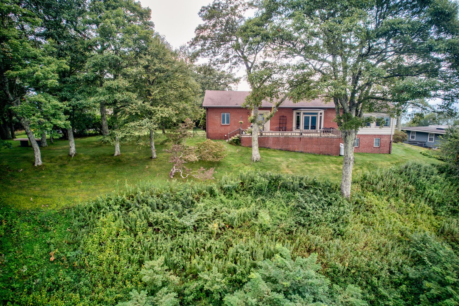 Une maison en briques avec un porche à l’avant, entourée d’arbres et d’une végétation luxuriante.