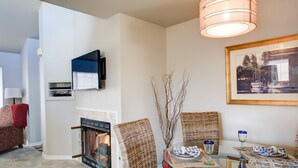 Un salon avec une cheminée, des fauteuils en osier, une table à manger avec des assiettes et des verres, un téléviseur fixé au mur et un tableau encadré accroché au mur.