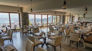 Restaurante com várias mesas e cadeiras, grandes janelas com vista para uma cidade e montanhas, e luminárias pendentes.