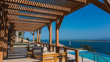 A covered outdoor seating area with wooden furniture and a pool overlooking the sea.