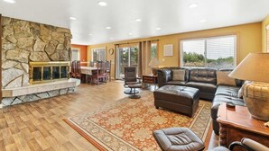 A spacious living room with a stone fireplace, leather sofas, and a large window providing natural light. - Pets OK, OCEAN VIEW, Beach Path, HOT TUB, Large Yard (FirstM) (Ocean Park)
