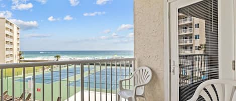 A balcony with a view of a tennis court and the ocean. There are two white chairs and a door with blinds.