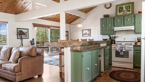 A kitchen with green cabinets, a white stove, and a dining area with a brown couch and a table. - LAKEFRONT - Boat Dock! Close to Slopes, HOT TUB! Game room! (Big Bear Lake)