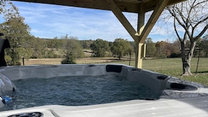 A hot tub under a wooden pergola with a view of a grassy field and trees.