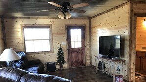 A living room with a leather couch, a TV on a wooden stand, a small Christmas tree, and a ceiling fan.