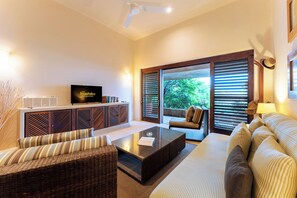 A modern living room with a flat-screen TV, wicker furniture, a glass coffee table, and a view of greenery through large windows. - Luxe Beach Villa in 5-star Oceanfront Community (Huatulco)