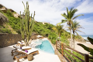 A poolside area with lounge chairs, a stone wall, and a view of the ocean. - Luxe Beach Villa in 5-star Oceanfront Community (Huatulco)