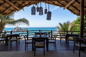 A wooden deck with tables and chairs overlooking a beach with a rocky outcrop and palm trees. - Luxe Beach Villa in 5-star Oceanfront Community (Huatulco)