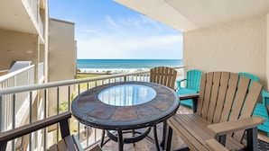 A balcony with a glass table and chairs, overlooking a beach with a clear view of the ocean.