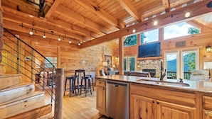 A spacious log cabin interior with a wooden staircase, a kitchen area, and a living room with a fireplace. - Charming 2 BR Log Home Super Views Paved Access Gravel Drive Hot Tub, & WIFI (Maggie Valley)