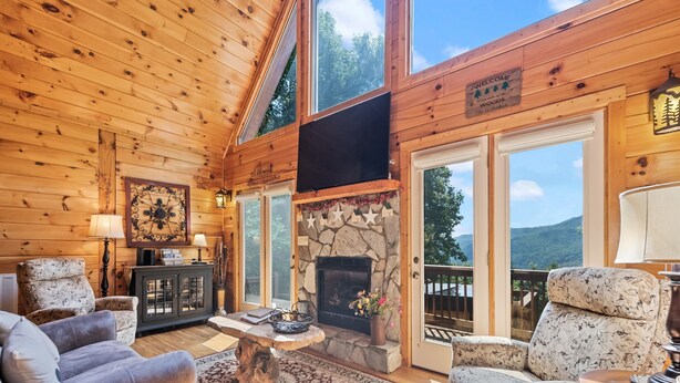 A cozy log cabin interior with a stone fireplace, wooden ceiling, and large windows offering scenic mountain views. - Charming 2 BR Log Home Super Views Paved Access Gravel Drive Hot Tub, & WIFI (Maggie Valley)