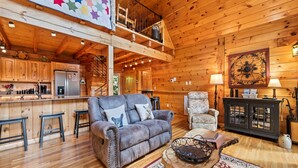 A cozy log cabin interior with a wooden bar, a gray sofa with a dog pillow, a patterned armchair, and a rustic coffee table. - Charming 2 BR Log Home Super Views Paved Access Gravel Drive Hot Tub, & WIFI (Maggie Valley)