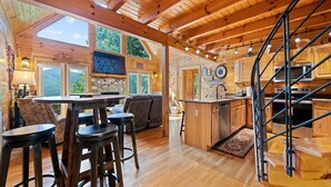 A wooden cabin interior with a dining area, kitchen, and a spiral staircase. - Charming 2 BR Log Home Super Views Paved Access Gravel Drive Hot Tub, & WIFI (Maggie Valley)
