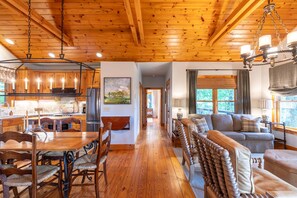 Una habitación amplia con techo de madera, un comedor con mesa y sillas de madera, y una sala de estar con sofá y vistas al exterior a través de amplios ventanales.
