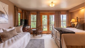 Una habitación acogedora con dos camas, un escritorio con televisor y ventanas amplias con vistas a árboles.
