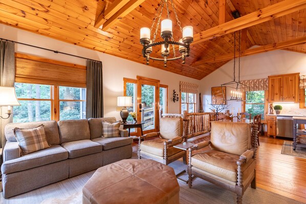 A cozy living room with a wooden ceiling, a sofa set, a dining table with chairs, and a kitchen area.