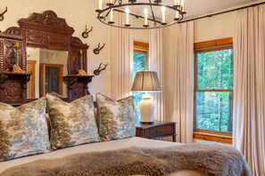 Un dormitorio con una cama grande, cabecera de madera, candelabro, espejo y ventana con cortinas.