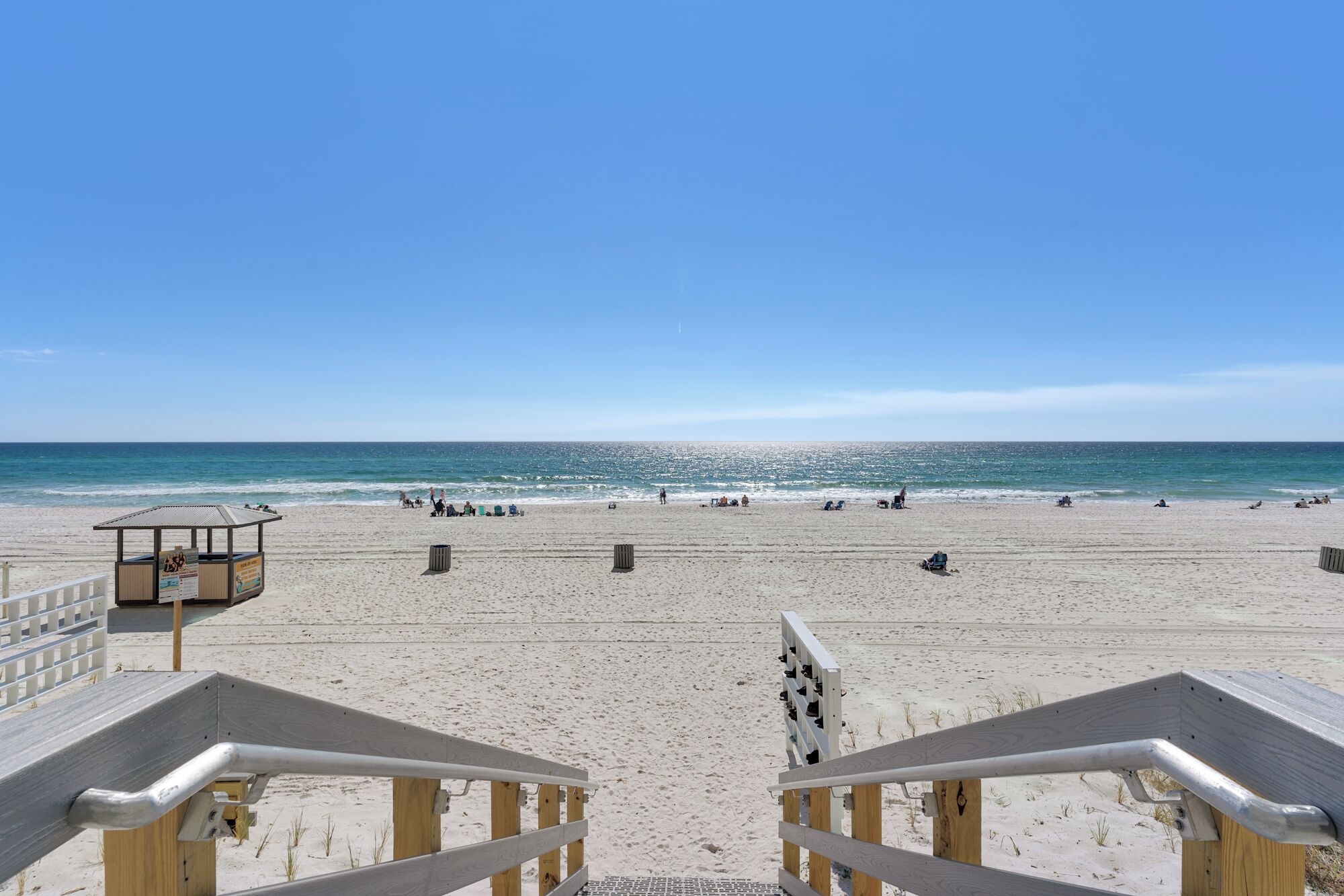 A beach with a wooden boardwalk leading to the water, a lifeguard station, and people relaxing on the sand.