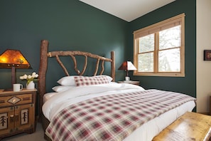 Une chambre à coucher avec une tête de lit en bois, une couvre-lit à carreaux, une table de chevet avec une lampe et une fenêtre donnant sur des arbres.