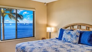 A bedroom with a large window overlooking the ocean, a bed with blue and white patterned bedding, a wooden headboard, and a nightstand with a lamp. - Oceanfront Kihei Beach Unit 410 Panoramic Views  Steps to the Beach! (Kihei)