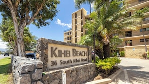 A sign for Kihei Beach at 36 South Kihei Road, with a stone wall and a multi-story building in the background. - Oceanfront Kihei Beach Unit 410 Panoramic Views  Steps to the Beach! (Kihei)