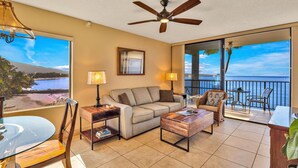 A spacious living room with a large window offering a view of the sea, a ceiling fan, and a balcony with outdoor furniture. - Oceanfront Kihei Beach Unit 410 Panoramic Views  Steps to the Beach! (Kihei)