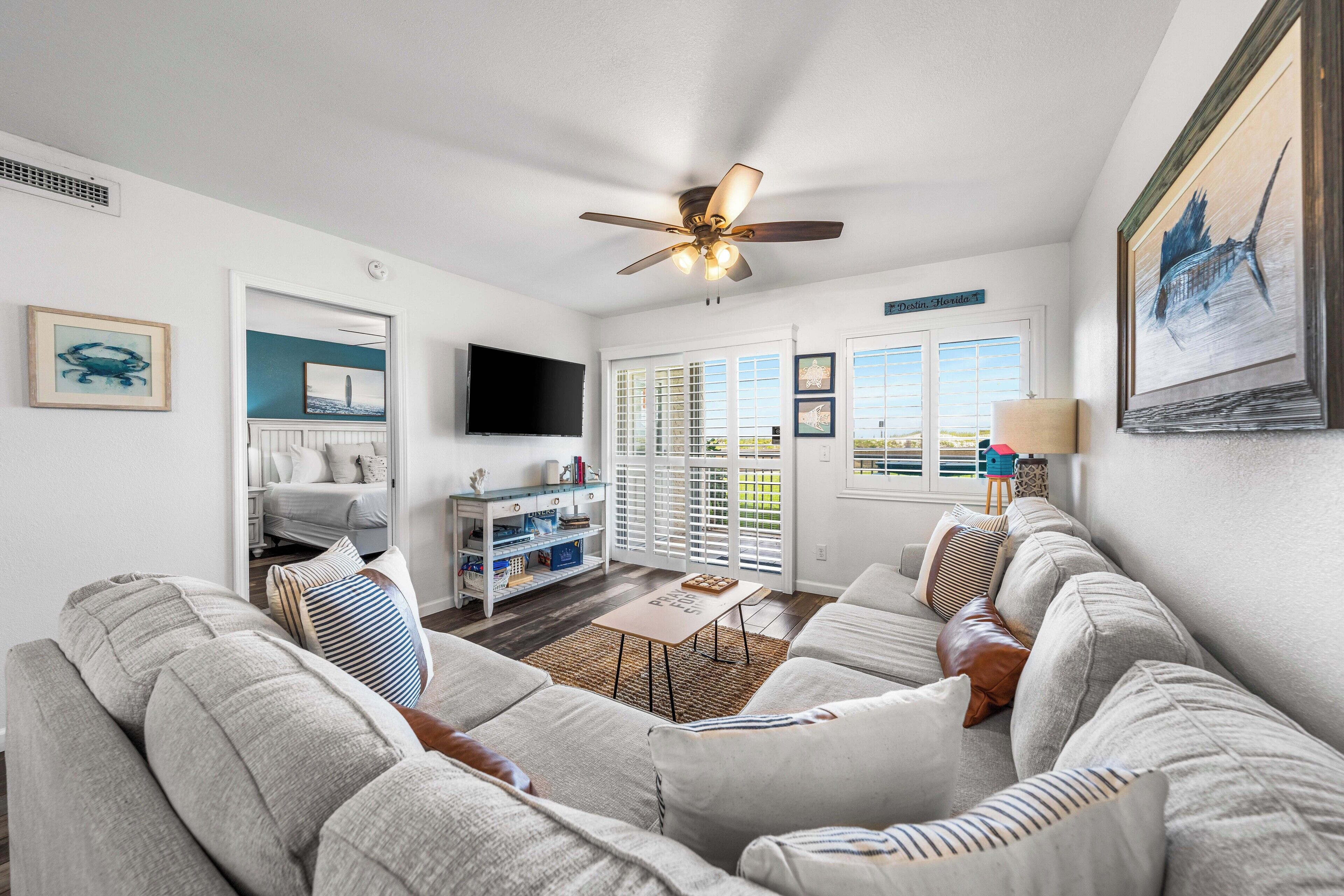A spacious living room with a large gray sectional sofa, a coffee table, and a ceiling fan. There is a TV mounted on the wall and a window with blinds.