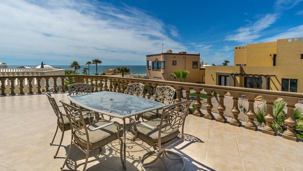 A terrace with a glass table and chairs, overlooking a beach and a clear blue sky.