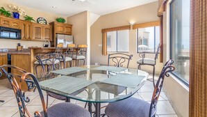 A modern kitchen with wooden cabinets, a microwave, and a refrigerator. There is a dining area with a glass table and metal chairs.